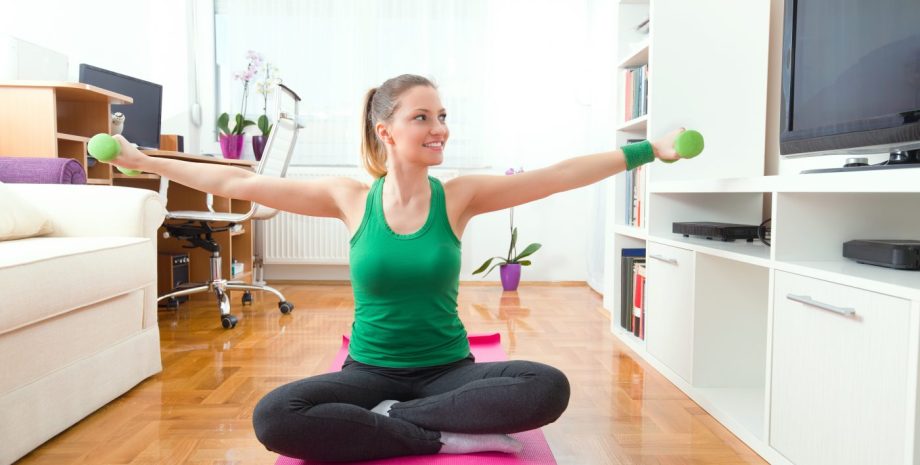 Happy young woman is exercising at home. She is sitting crossed-legged on the mat and doing exercise for her arms with a pair of green hand weights.