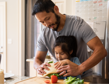 A father leaning over their child's shoulders helping them make a sandwich.